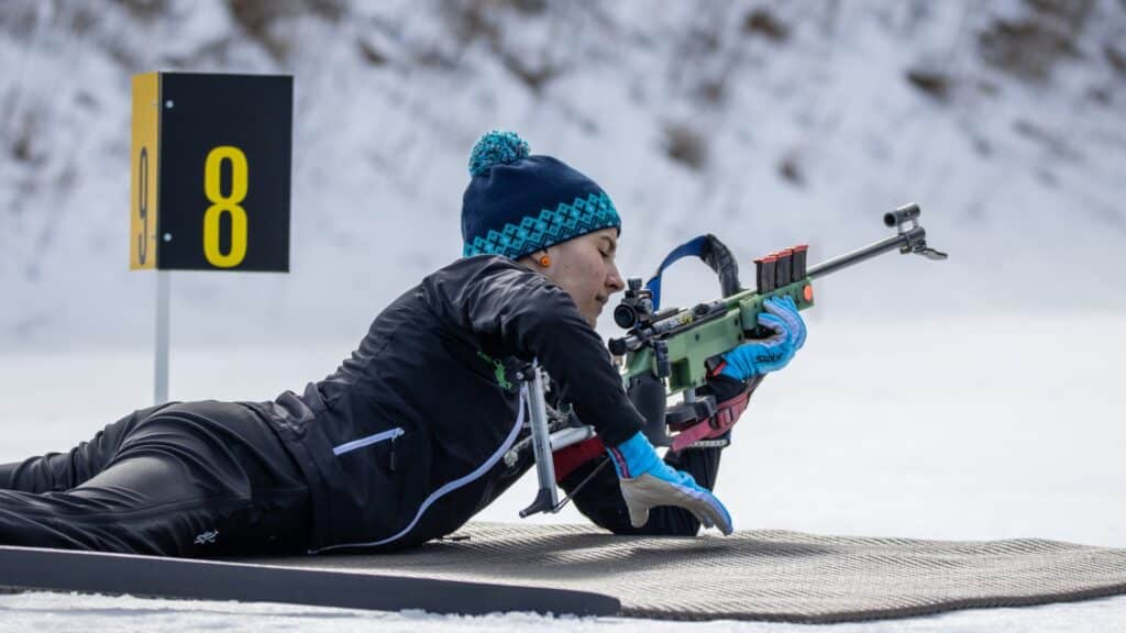 Biathlon Frau liegt am Boden und hält ein Gewehr in der Hand, verschneiter Hintergrund